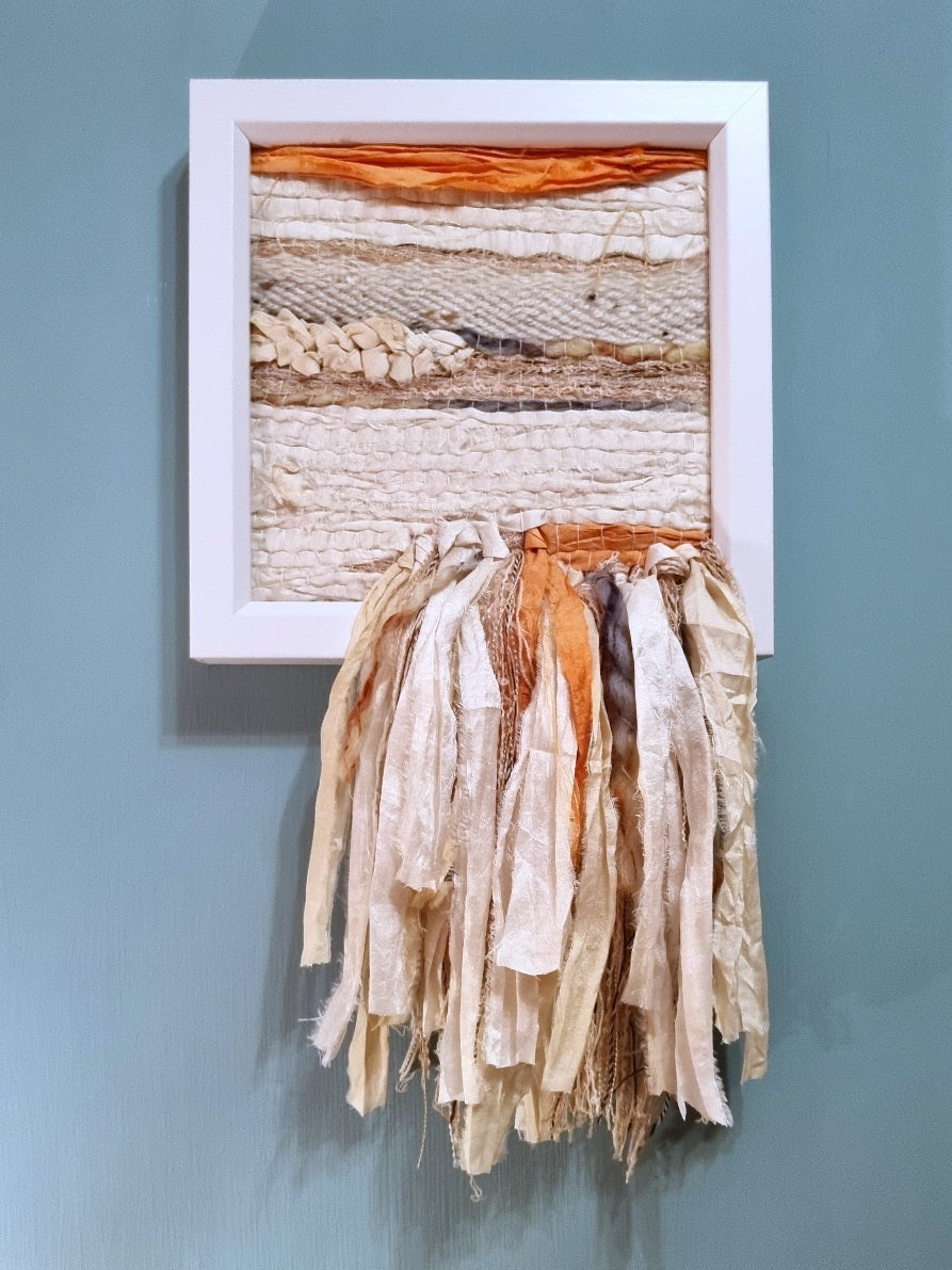 Hand-woven wall hanging in soft off-white recycled silk, with coral and sea blue details. A fringe cascades down from the woven piece, hanging below the white wooden box frame.