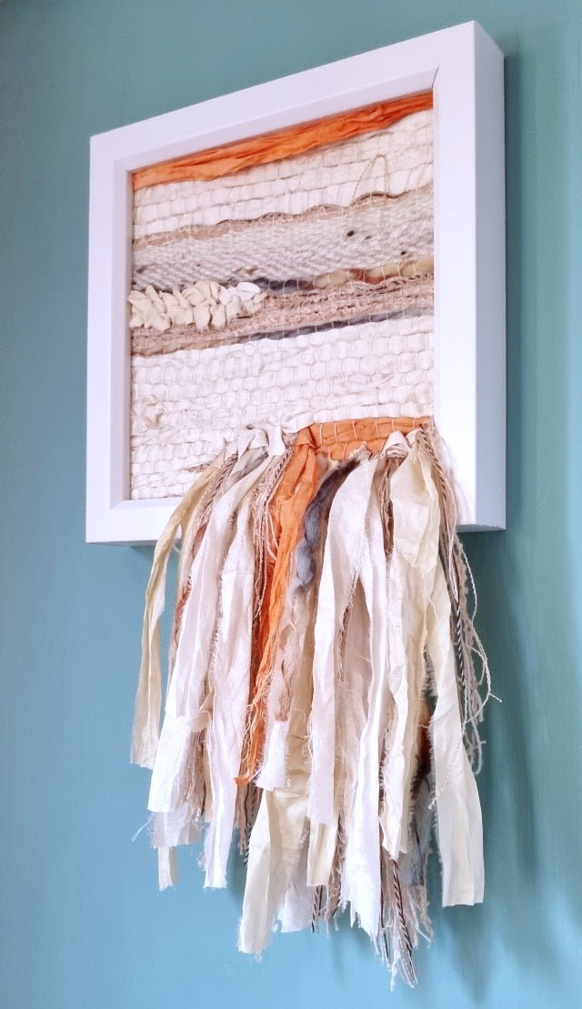 Hand-woven wall hanging in soft off-white recycled silk, with coral and sea blue details. A fringe cascades down from the woven piece, hanging below the white wooden box frame.